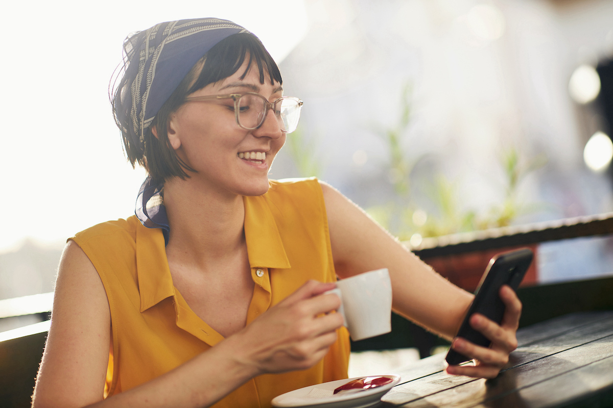 Young woman drinking coffee on a patio and smiling at her smartphone enjoying by the benefits of outsourcing IT support to an MSP