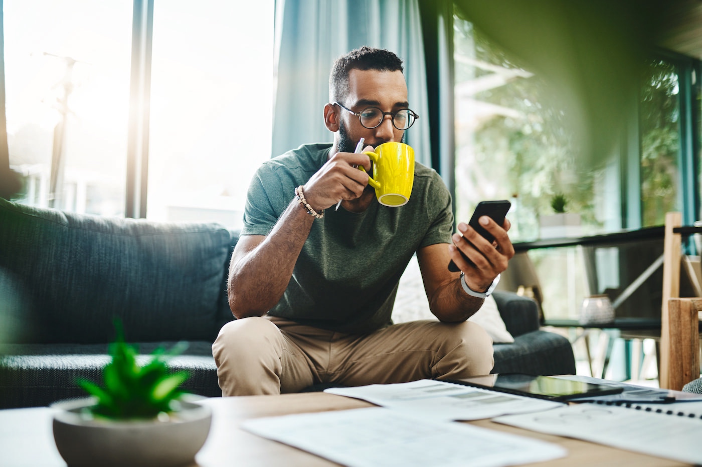 Young man in his living room drinking coffee, looking at his smartphone and paperwork, and planning how to start an MSP